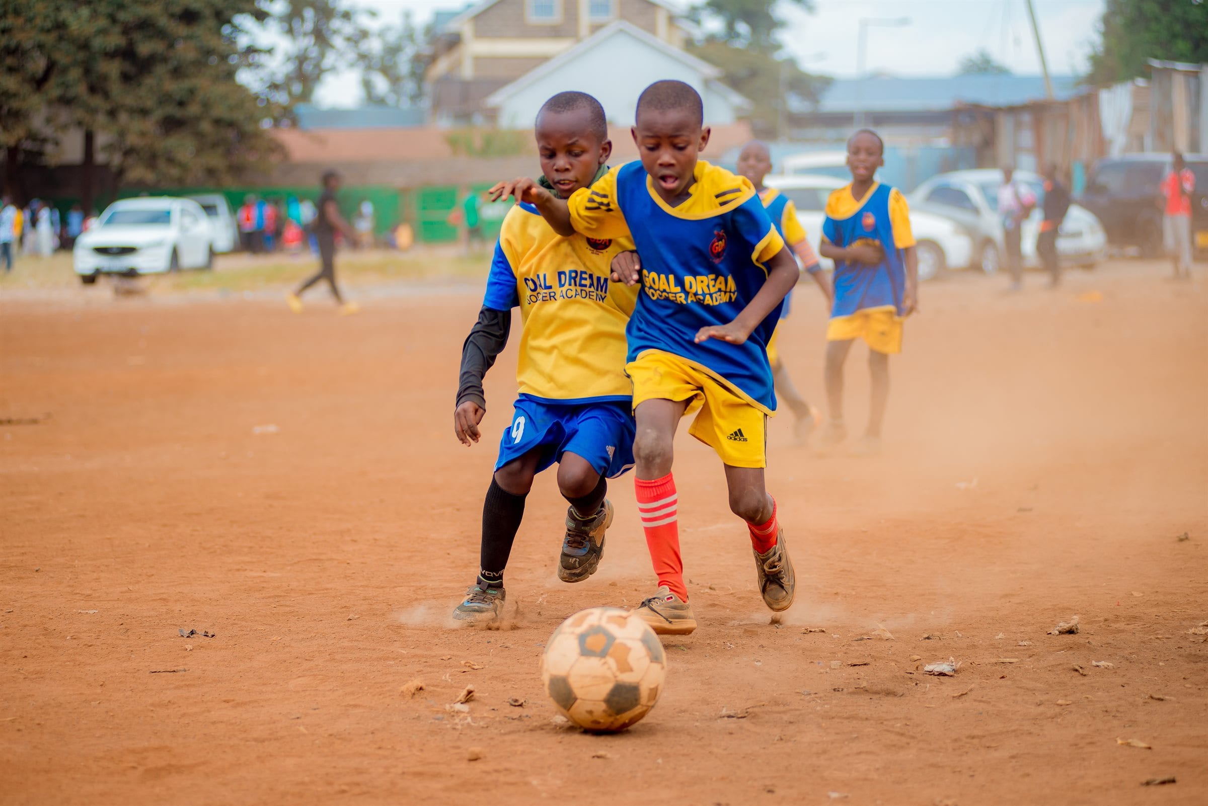 Two Goal Dream Soccer Academy players, one in a yellow bib and one in blue, run shoulder to shoulder chasing the ball across the dirt pitch at Baba Dogo