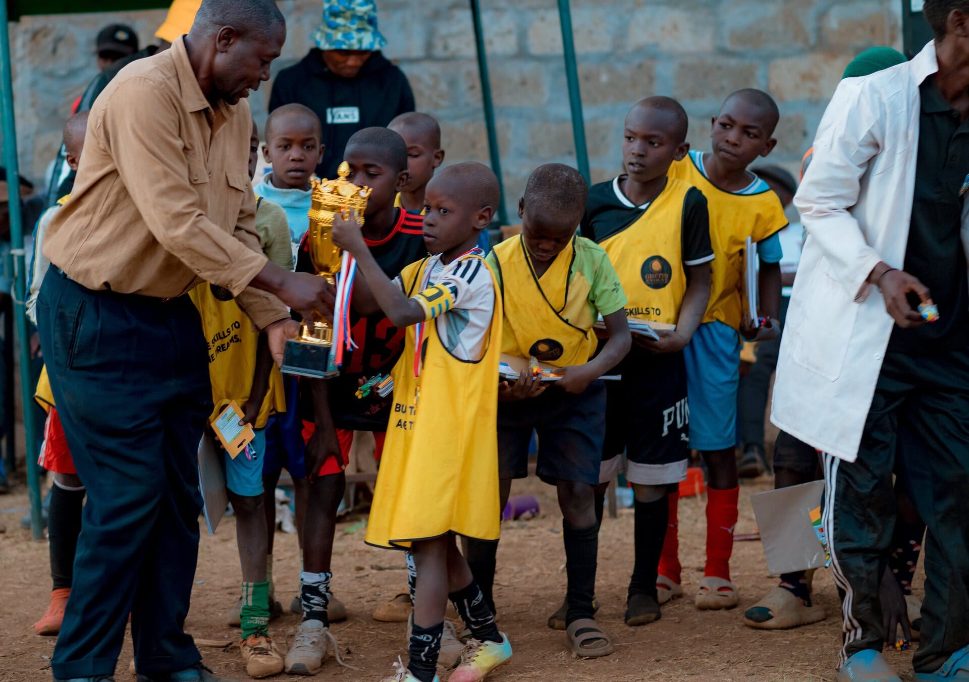 An adult hands a gold trophy to a boy in a yellow bib while other children wait alongside him holding their certificates