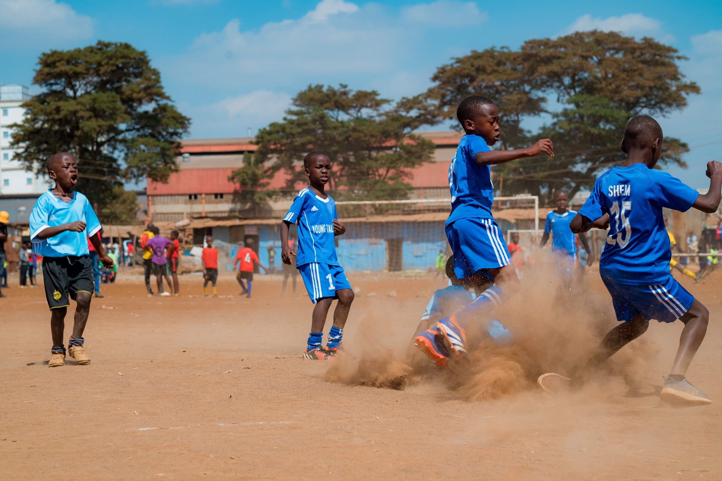 Young Goal Dream players in blue kit turn and sprint through a cloud of dust they have kicked up on the dry pitch, with trees and rooftops behind them