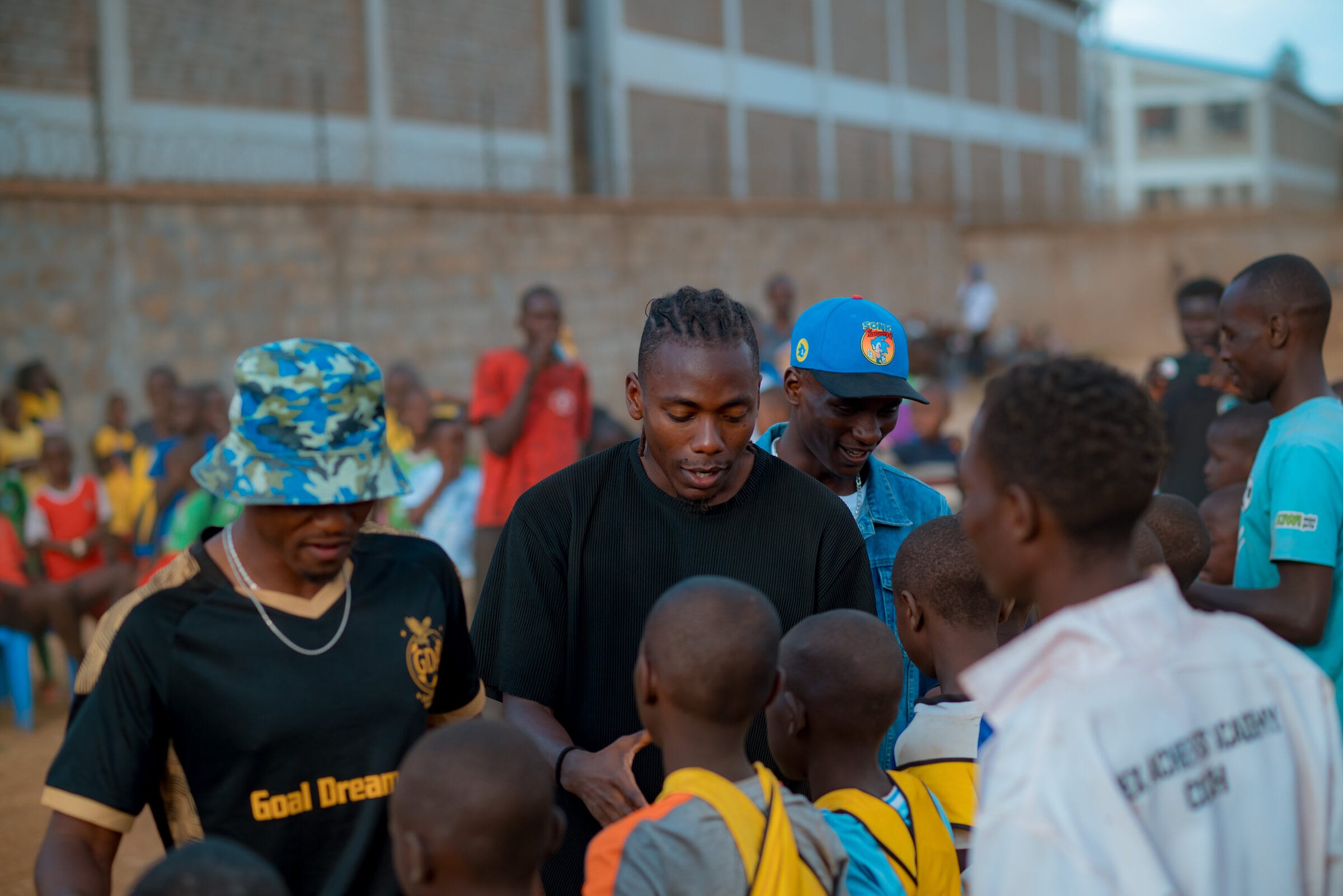 Goal Dream team members, one in a black shirt carrying the Goal Dream name, lean in to greet a group of children at the edge of the pitch