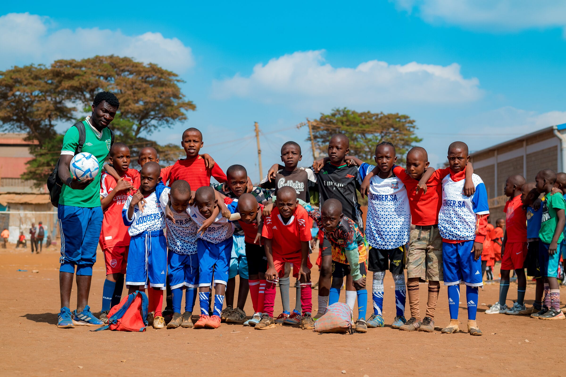 A youth team stands arm in arm across the pitch beside their coach, who holds a football under one arm, under an open sky