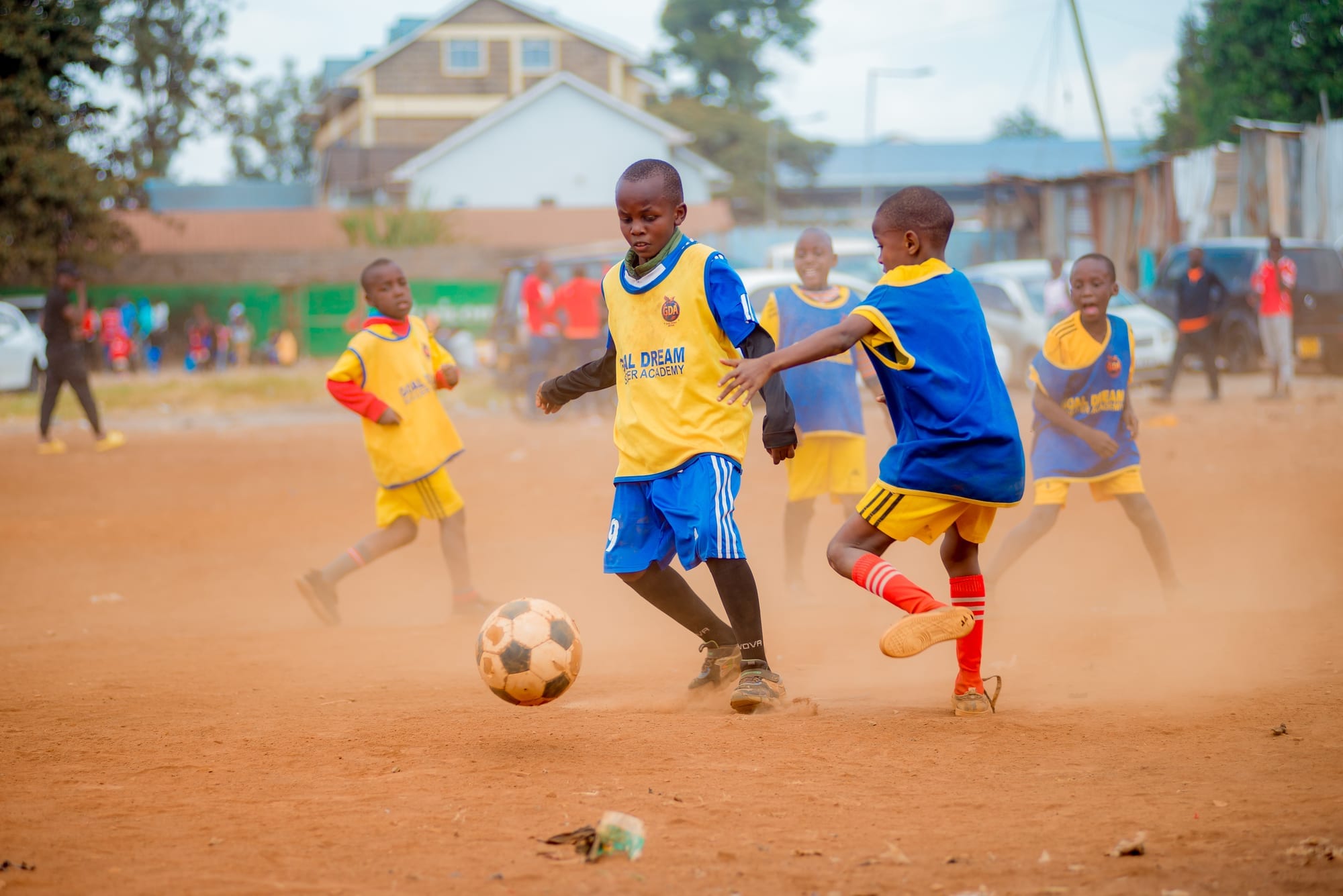 A Goal Dream Soccer Academy player in a yellow training bib drives the ball forward on the dirt pitch as an opponent in blue closes him down, with other players and parked cars behind
