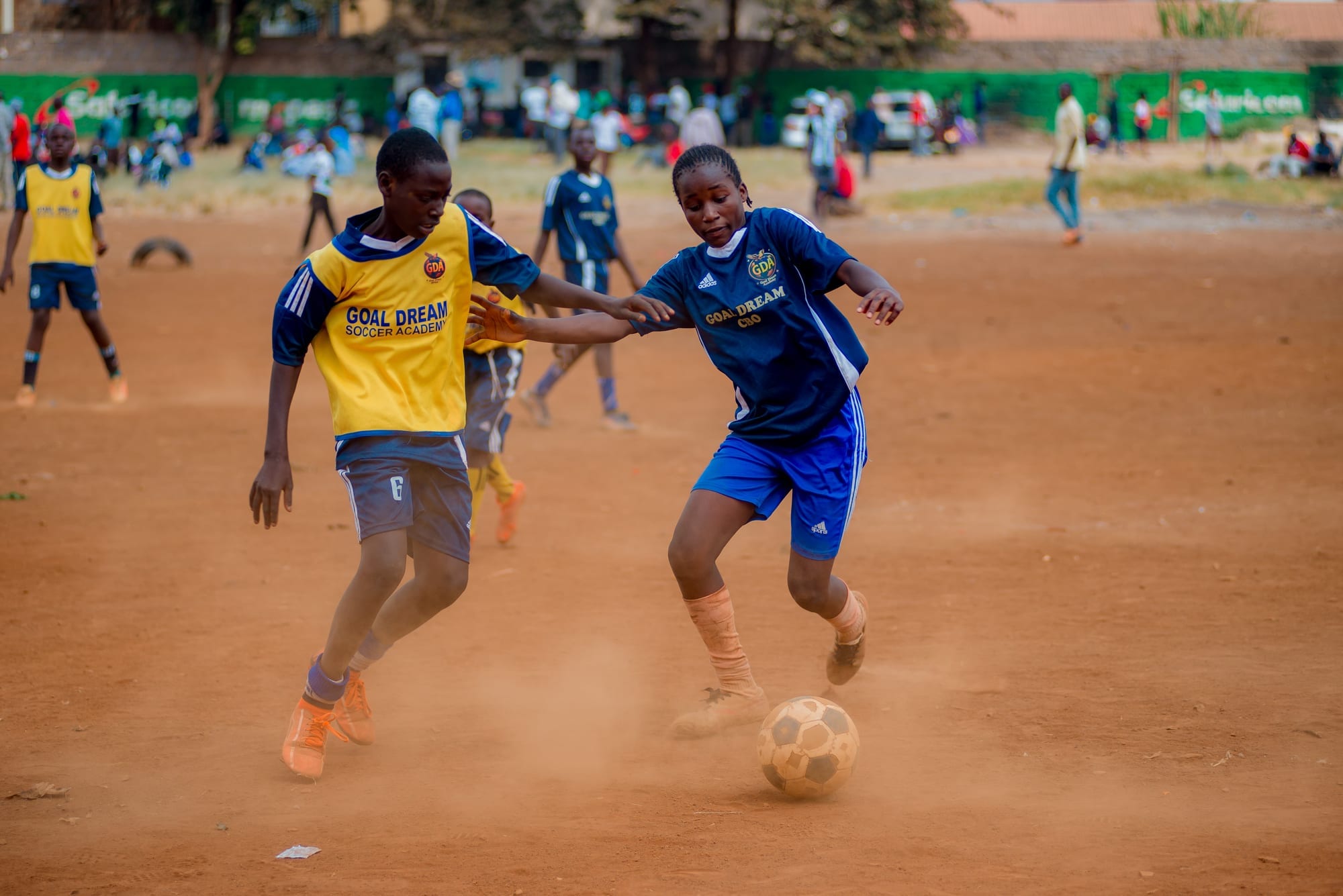 A girl in a navy Goal Dream shirt runs the ball past a boy in a yellow Goal Dream bib during a match on the dirt pitch, with spectators along the touchline behind