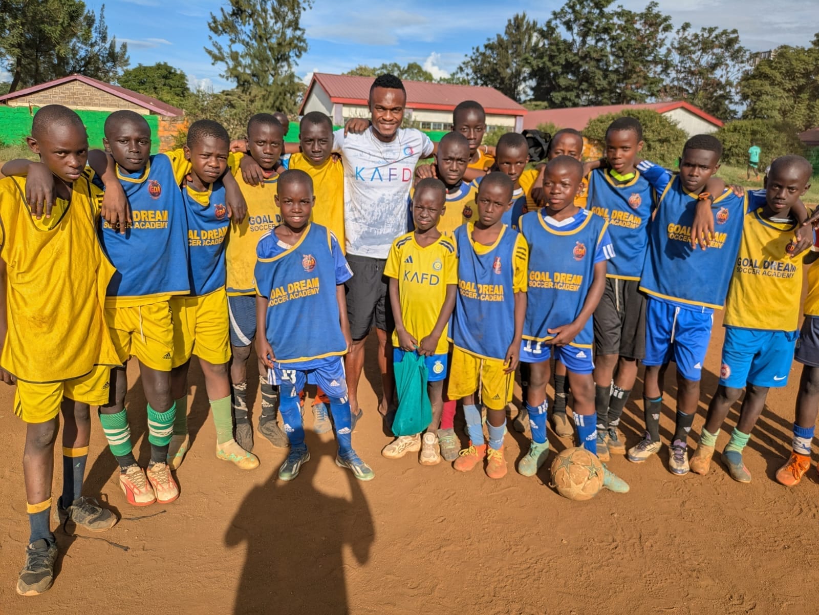 A Goal Dream coach stands in the middle of a long line of young players in Goal Dream bibs, arms around one another's shoulders, on the pitch with buildings and trees behind